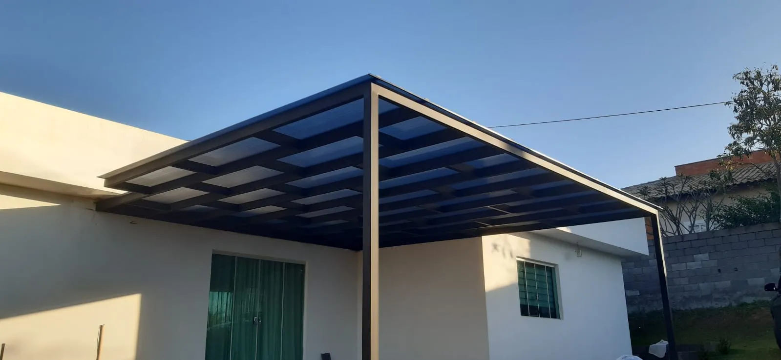 Pergolado de madeira visto de baixo, com céu azul e casa moderna ao fundo
